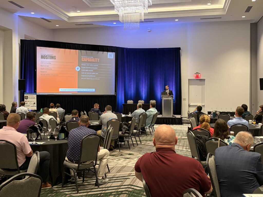 Dr. Kollin Napier delivers the enHance keynote from a podium before a seated audience as a projected slide contrasts hosting AI infrastructure with building statewide capability