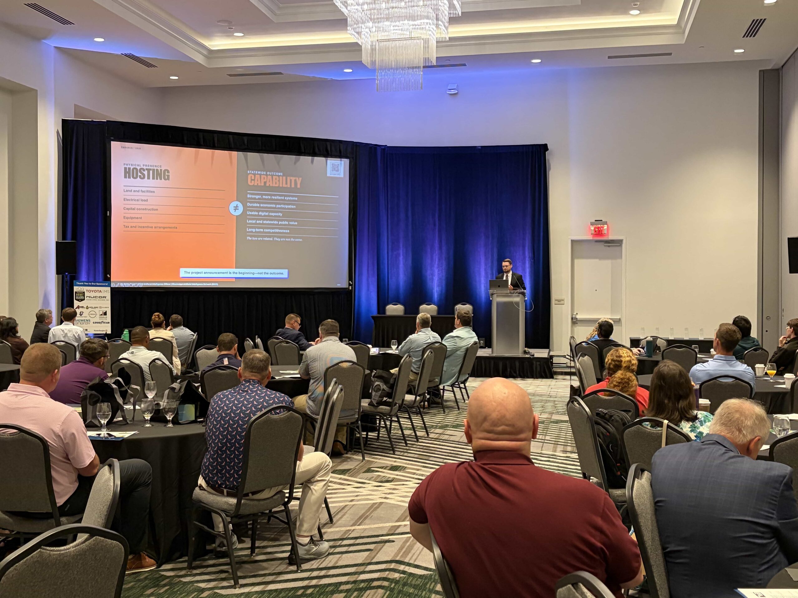 Dr. Kollin Napier delivers the enHance keynote from a podium before a seated audience as a projected slide contrasts hosting AI infrastructure with building statewide capability