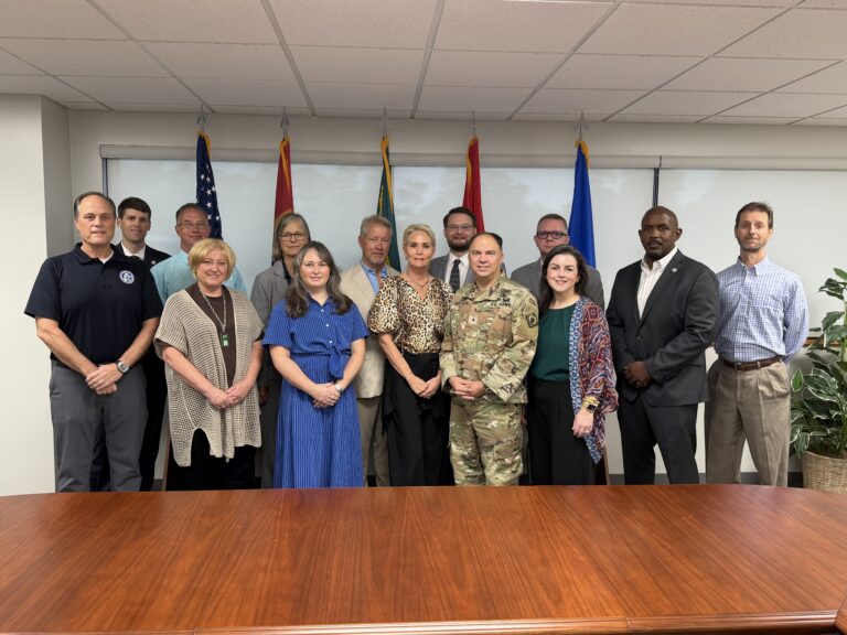 Task force members and invited speakers stand together in a meeting room at Camp Shelby.