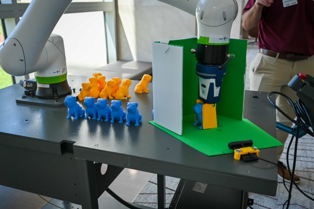 Collaborative robotic arms work with blue and orange model pieces during an automated manufacturing demonstration.