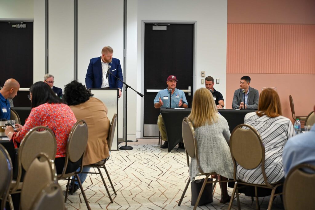 A moderator stands at a podium beside panelists seated at microphones during AI in Manufacturing Day.