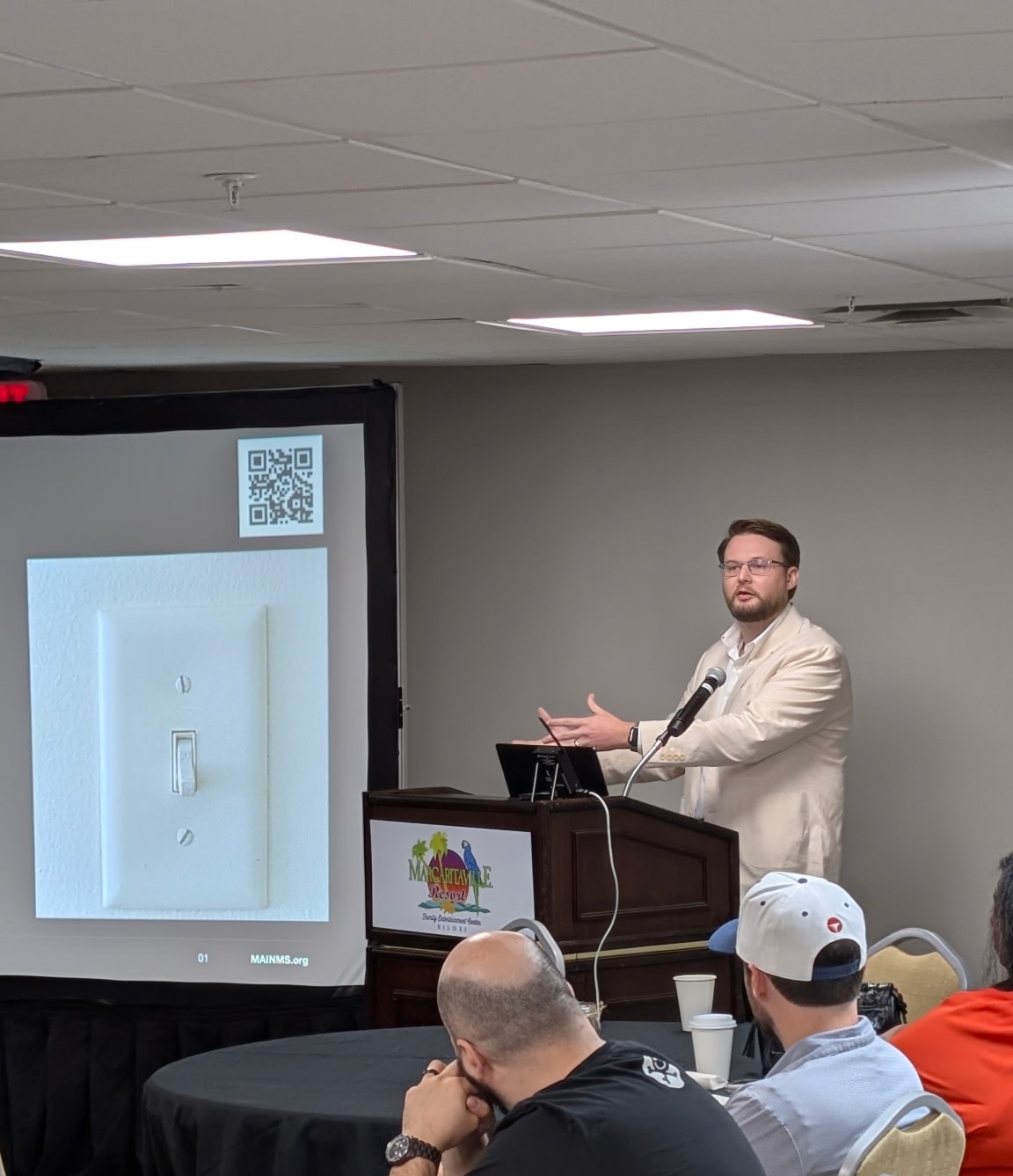 Dr. Kollin Napier speaks at a podium beside a projected image of a light switch, with attendees in the foreground.