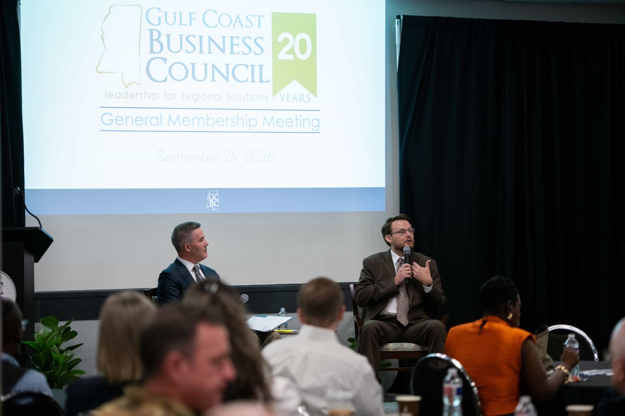 Dr. Kollin Napier and Jamie Miller onstage beneath the Gulf Coast Business Council meeting display.