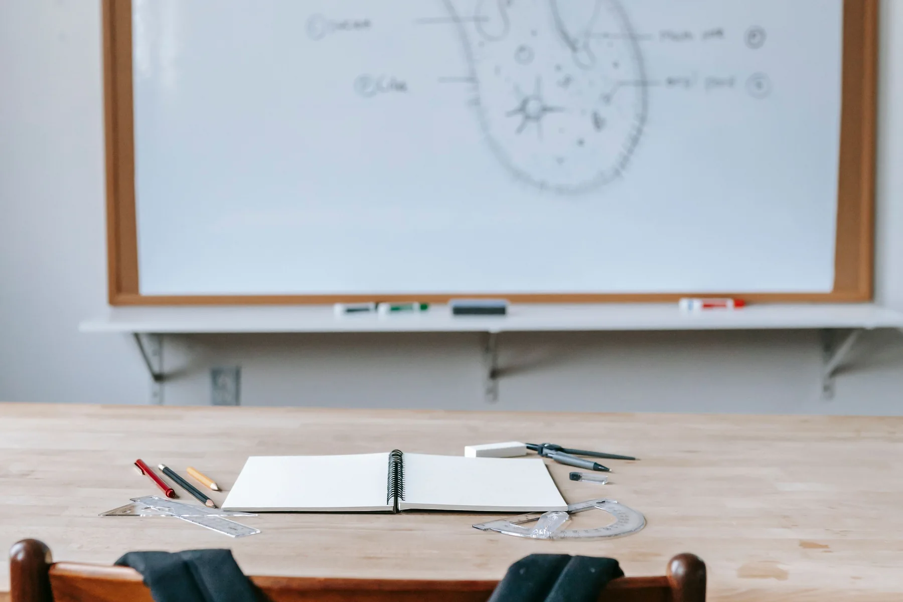 An unoccupied classroom desk with an open notebook and measuring tools, with a whiteboard behind it.