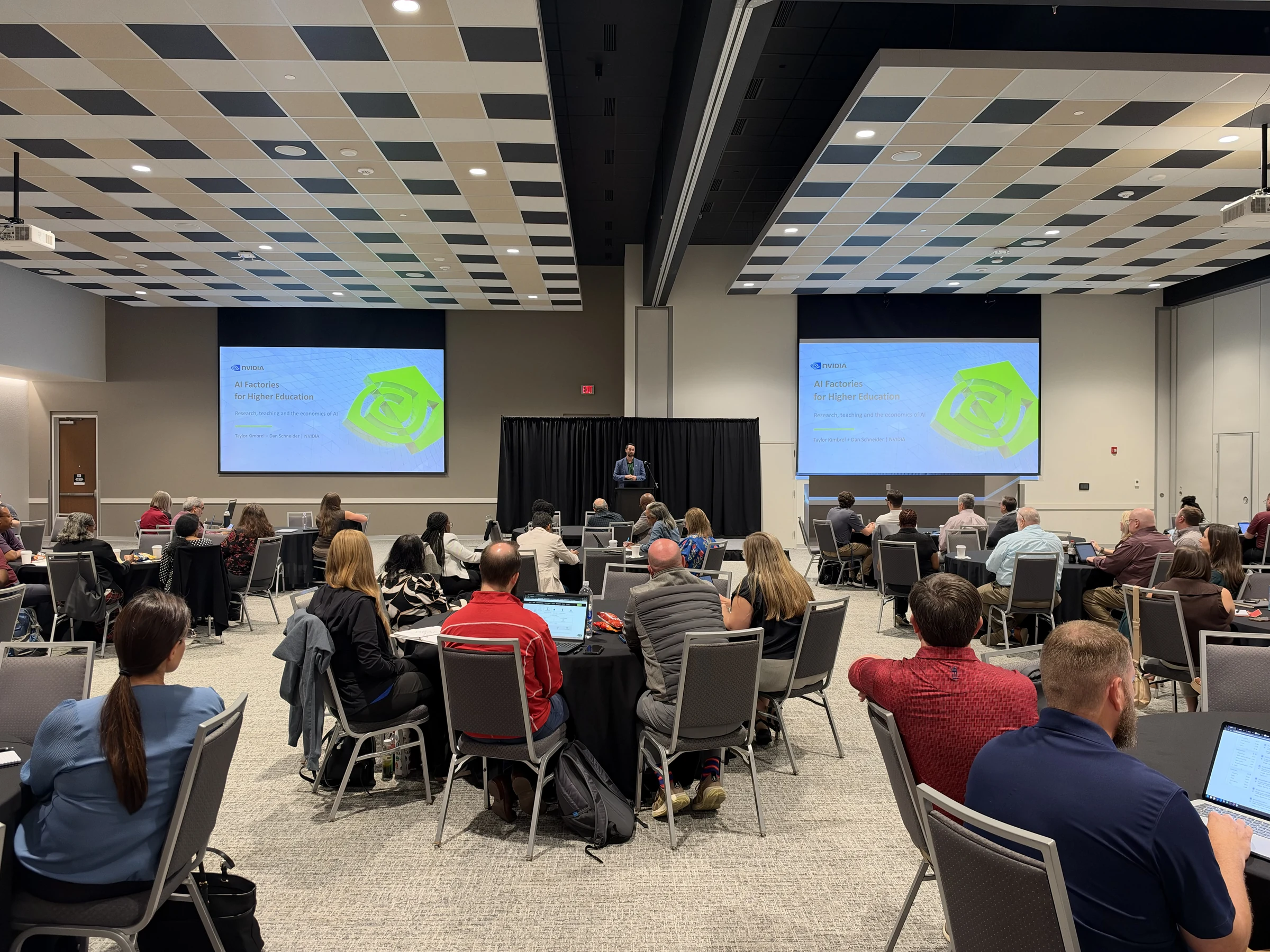 Speaker at a lectern between screens reading AI Factories for Higher Education, with workshop attendees seated in the foreground.