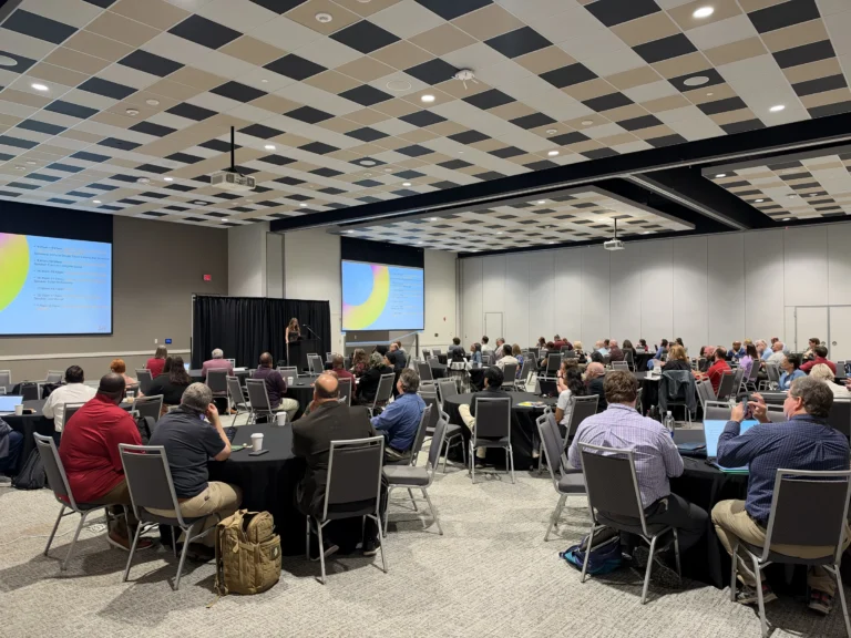 Attendees seated at tables face a speaker and agenda screens during the AI executive workshop at The Mill at Mississippi State University.