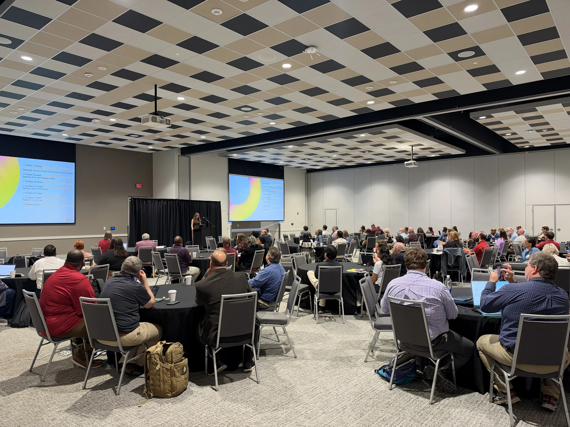 Attendees seated at tables face a speaker and agenda screens during the AI executive workshop at The Mill at Mississippi State University.