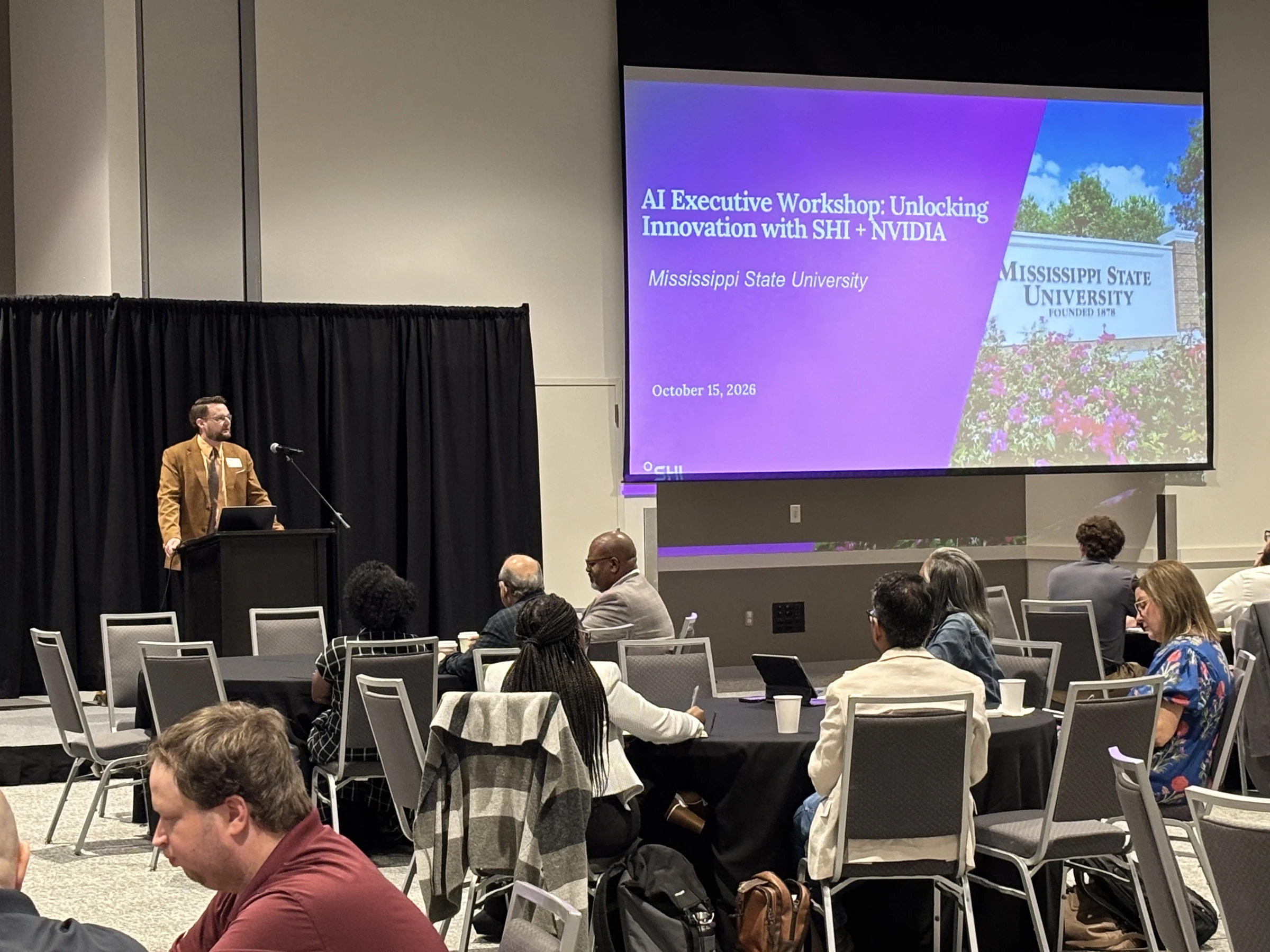 Speaker at a lectern beside a screen displaying AI Executive Workshop: Unlocking Innovation with SHI + NVIDIA and the October 15, 2026 date.